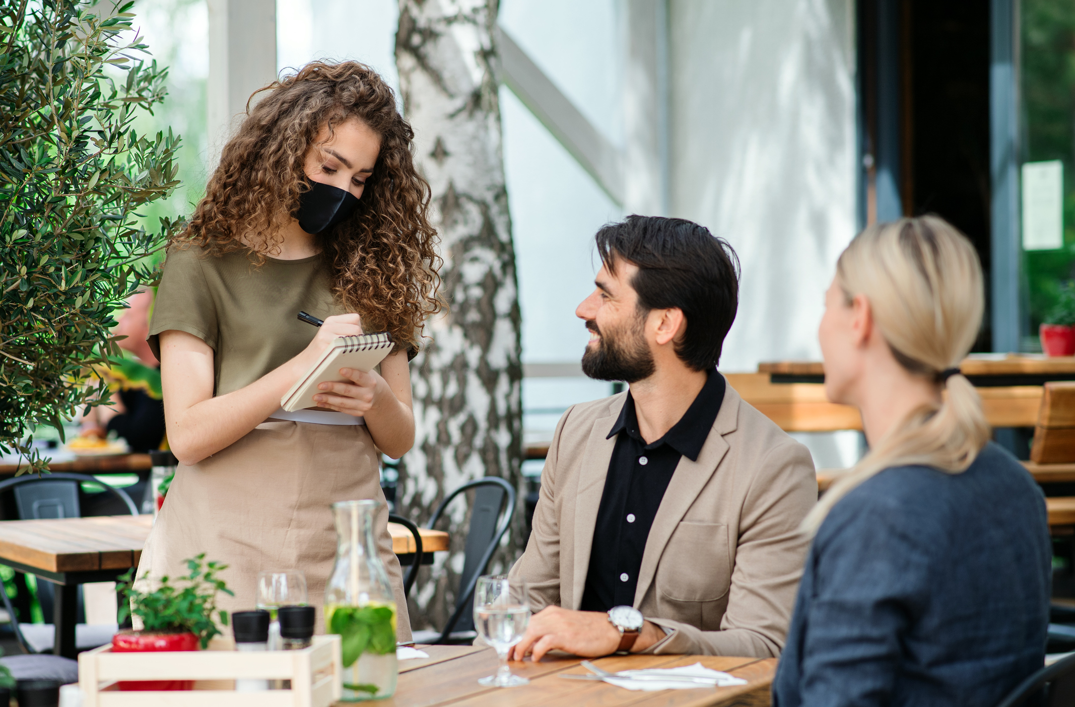 Waitress with face mask serving happy couple outdoors on terrace restaurant.